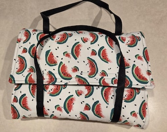 May include: A white picnic blanket with a watermelon slice pattern in red and green, with black seeds. The blanket has a black handle and straps for carrying. The blanket is folded and secured.