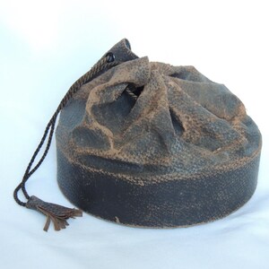 May include: A round, dark brown leather drawstring pouch with a textured surface and a matching tassel. The pouch has a lighter brown, distressed appearance on the top, and a dark brown drawstring.