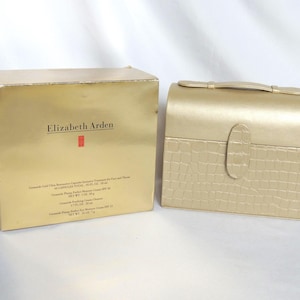May include: A gold-toned gift set by Elizabeth Arden, featuring a box and a matching cosmetic case. The box has the brand name and product information printed on it. The cosmetic case has a handle and a textured, crocodile-like pattern.
