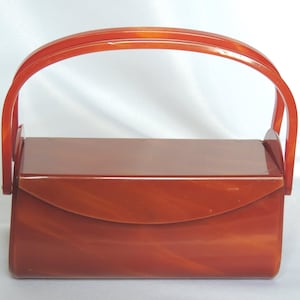 Pode incluir: Bolsa de mão retangular vintage, em tom castanho-avermelhado quente, com alça curva e fecho de aba. A bolsa tem um acabamento brilhante. A alça é arqueada e presa na parte superior da bolsa.