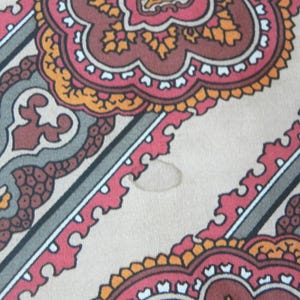 May include: Close-up of a patterned fabric with a beige background. The design features repeating floral and geometric shapes in red, brown, orange, and grey. Diagonal stripes with white borders intersect the pattern.