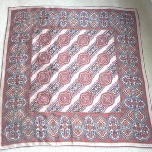 May include: A square scarf with an intricate design. The scarf has a border with ornate shapes in red, grey, and cream. The centre features diagonal stripes with floral-like patterns in similar colours. The scarf is likely made of silk or a similar fabric.