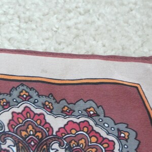 May include: A square textile with a detailed paisley pattern. The border is a light beige, framing a deep burgundy field with orange, grey, and red floral motifs. Likely a scarf or decorative cloth, with a classic design.