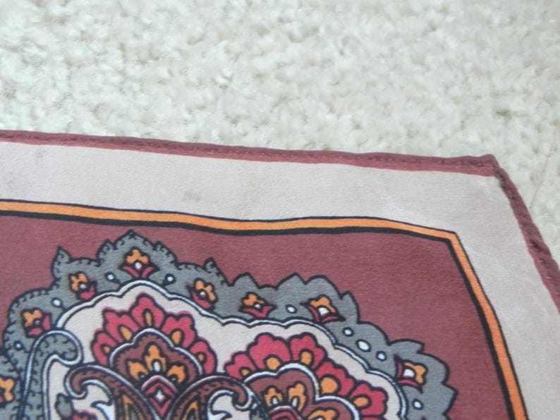 May include: A square textile with a detailed paisley pattern. The border is a light beige, framing a deep burgundy field with orange, grey, and red floral motifs. Likely a scarf or decorative cloth, with a classic design.