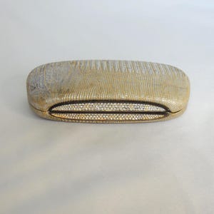 Puede incluir: Un estuche rectangular dorado para gafas con un patrón texturizado similar al de un reptil. El estuche tiene una parte superior e inferior redondeadas con un ribete negro y un patrón decorativo en forma de diamante alrededor de la abertura.