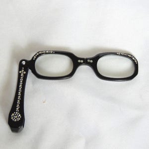 May include: Black reading glasses with a folding arm, adorned with small, sparkling embellishments. The frames are oval-shaped and feature decorative accents. The glasses are set against a white background.