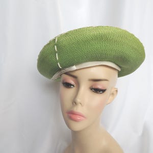May include: A green woven hat with a white interior and decorative white stitching. The hat is a beret style and is displayed on a mannequin head. The hat is a light green color.