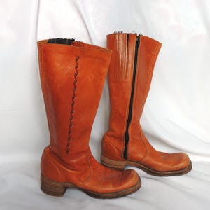 Puede incluir: Un par de botas altas de cuero naranja. Una bota tiene un diseño cosido decorativo, mientras que la otra presenta una cremallera negra. Las botas tienen una apariencia vintage desgastada con tacones de madera.