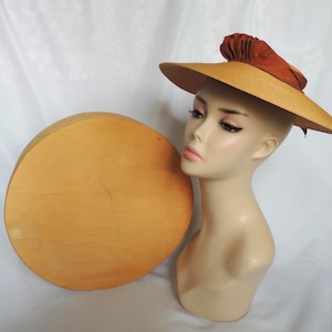 May include: A vintage sun hat with a wide brim and a decorative accent. The hat is displayed on a mannequin head next to a round wooden hat box. The hat features a light brown brim and a dark brown accent.