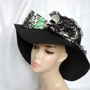 May include: Black wide-brimmed hat with a decorative bow. The bow features a black and white patterned fabric with a green and red design, and is trimmed with black lace. The hat is displayed on a mannequin.