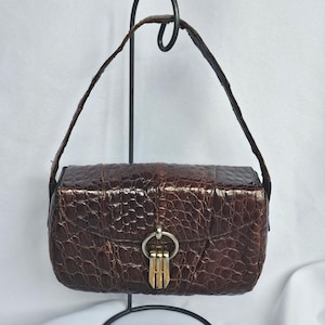 Puede incluir: Un bolso de mano rectangular marrón con un patrón texturizado similar a la piel de cocodrilo. El bolso tiene un asa corta y un cierre dorado. El bolso se exhibe sobre un soporte de metal negro sobre un fondo blanco.