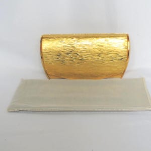 Puede incluir: Un clutch dorado con una superficie texturizada y bordes redondeados. El clutch es rectangular y tiene una funda protectora suave y de color claro a juego. El clutch es probablemente para eventos formales.