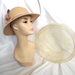 May include: A tan fedora-style hat with a wide brim and a decorative feather and flower accent. The hat is displayed on a mannequin head. A clear, round, decorative plate is next to the mannequin.