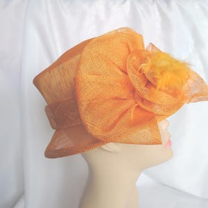 May include: An orange hat with a large, decorative bow and feather accents. The hat is made of a textured fabric and is displayed on a mannequin head. The hat has a wide brim and a band around the crown.
