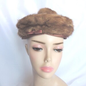 May include: A vintage brown fur hat with a small bow detail. The hat is a light brown color and has a rounded shape. The hat is displayed on a mannequin head with a pale complexion and pink lipstick.