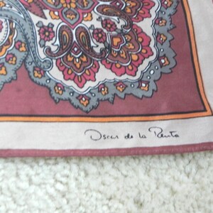 May include: A silk scarf featuring a paisley pattern in red, orange, grey, and cream. It has a burgundy border and the signature "Oscar de la Renta" in the corner.