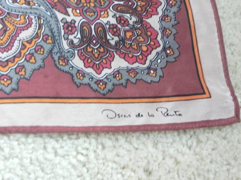 May include: A silk scarf featuring a paisley pattern in red, orange, grey, and cream. It has a burgundy border and the signature "Oscar de la Renta" in the corner.