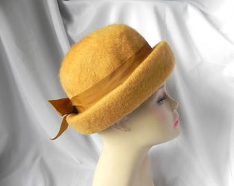 May include: Mustard yellow cloche hat with a matching ribbon band and bow. The hat has a rounded crown and a slightly turned-up brim. A vintage-inspired accessory.