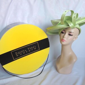 May include: A pale green hat with ribbon detailing and embellishments is displayed on a mannequin head. A yellow hat box with a black band and the text "Donna Vinci Couture" is also visible.