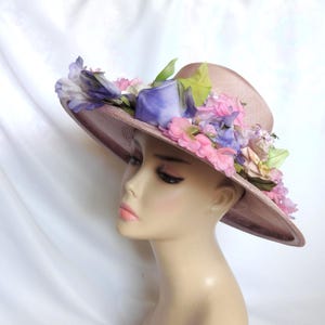 May include: A wide-brimmed, mauve-colored hat adorned with an array of colorful silk flowers. The floral arrangement includes pink, purple, and blue blossoms, with green leaves. The hat is displayed on a mannequin head against a white backdrop.