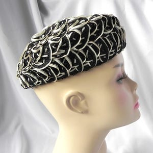 May include: A black and white hat with a woven design. The hat is a beret style, with a rounded crown and a close-fitting brim. The hat is made of a black material with white ribbon woven throughout.