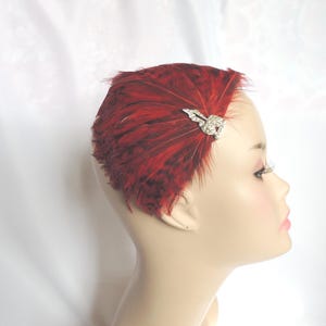May include: A red feather headpiece with a decorative rhinestone clip. The headpiece is made of layered red feathers and is attached to a small clip. The headpiece is worn on a mannequin head.