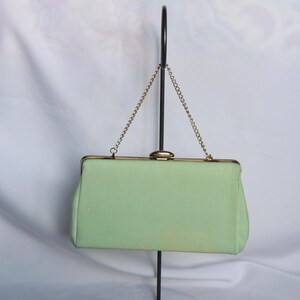 Op de afbeelding: Een lichtgroene rechthoekige clutch met een goudkleurige sluiting en kettingriem. De tas is gemaakt van een textuurstof en wordt getoond tegen een witte achtergrond.
