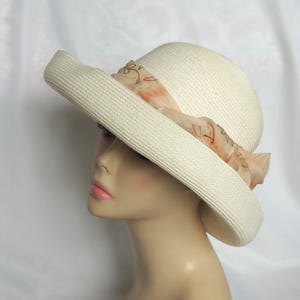 May include: A cream-colored straw hat with a wide brim and a rounded crown. The hat is adorned with a soft, peach-toned fabric band wrapped around the base, featuring a delicate, thin brown line pattern printed across the fabric material.