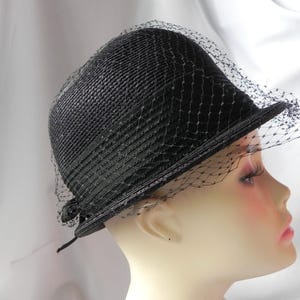 May include: A black hat with a structured brim and a delicate veil. The hat is made of a woven material, and the veil is attached to the brim. The hat is displayed on a mannequin head.