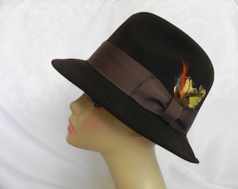 May include: A dark brown fedora hat with a brown ribbon band and a decorative feather accent. The hat is displayed on a mannequin head, showcasing its shape and style. The feather accent includes orange, yellow, and blue feathers.