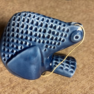 May include: A blue ceramic fish figurine with a textured, scale-like pattern. The fish has a small eye and a string attached near the top. The figurine is a decorative item.
