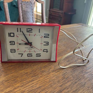 May include: A vintage General Electric alarm clock with a red frame and a white face. The clock features black numerals, red hands, and a beige power cord. The clock is on a wooden surface.