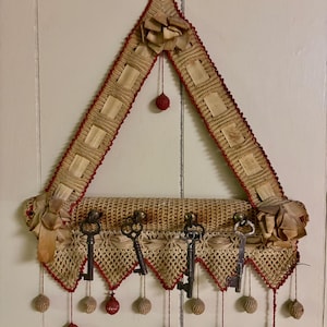 Puede incluir: Un llavero hecho a mano con un diseño triangular. El llavero está hecho de tela de ganchillo beige con ribetes rojos y detalles decorativos. Cuenta con varias llaves antiguas colgando de la parte inferior y pompones decorativos rojos y beige.