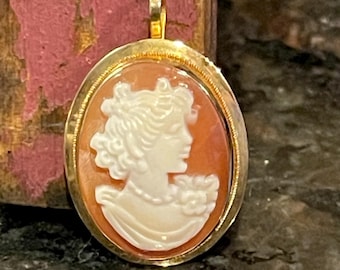 Puede incluir: Colgante ovalado de oro con un camafeo que muestra el perfil de una mujer en blanco sobre un fondo naranja. El colgante tiene un marco dorado y un lazo para una cadena. El camafeo representa a una mujer con cabello rizado y un escote drapeado.