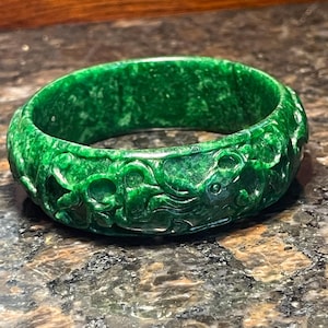 May include: A vibrant green jade bangle bracelet. The bracelet features intricate carvings along its surface, showcasing a detailed design. The polished jade reflects light, highlighting the rich green color and the carved details. This piece is a statement accessory.