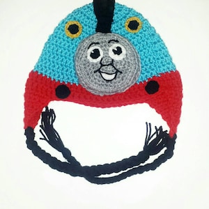 May include: A blue and red crocheted hat with a black crocheted tassel. The hat features a grey crocheted face with black eyes and a white smile. The face is reminiscent of a cartoon train engine.
