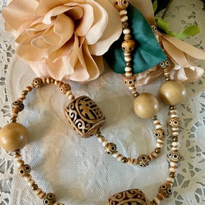 May include: A long, double-strand necklace made of wooden beads. The beads are carved with intricate designs and some are round. The necklace is beige and brown in color.
