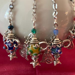 May include: Close-up of several decorative teapot charms with colorful glass beads and silver-toned metal accents. Each charm features a miniature teapot design with intricate details and a small bead at the top. The charms are suspended from a silver chain.