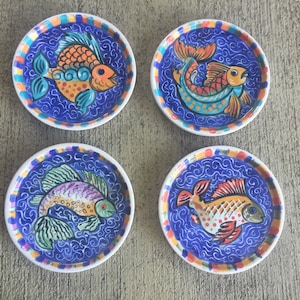 May include: Set of four hand-painted ceramic coasters. Each coaster features a unique, colorful fish design on a blue background. The fish are orange, yellow, green, and purple, with white accents. The coasters have a white border with a colorful trim.