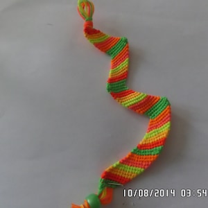 May include: A multi-colored friendship bracelet with green, orange, and yellow stripes. The bracelet is tied with a knot at the top and has a green bead at the bottom.