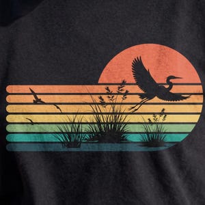 May include: A black t-shirt featuring a graphic print of a bird in flight over marsh grass. The design includes a sunset motif composed of horizontal stripes in shades of orange, yellow, green, and blue, creating a retro-inspired landscape scene.
