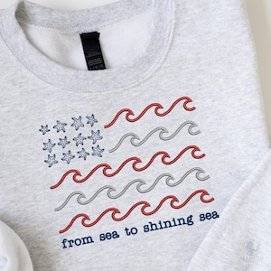 May include: Light gray sweatshirt with an embroidered design of the American flag, featuring blue starfish in the corner and red and gray waves. The text "from sea to shining sea" is below the flag.