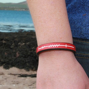 Puede incluir: Pulsera de cuero rojo y marrón con costuras blancas y una franja blanca. La pulsera se lleva en la muñeca. El fondo muestra una playa, agua y un cielo azul.