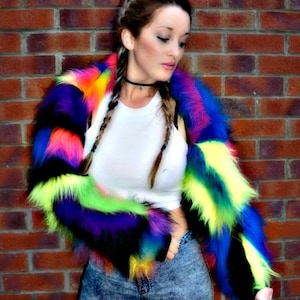 Può includere: Una donna indossa una giacca di pelliccia sintetica colorata con una canotta bianca e jeans blu. La giacca ha strisce di pelliccia sintetica rosa, gialla, verde, blu e nera.