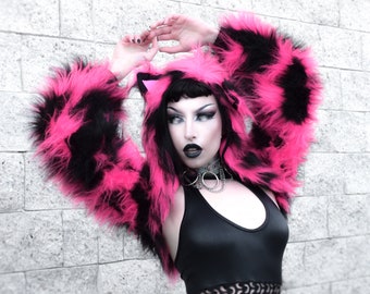 May include: A pink and black faux fur jacket with a cat ear hood. The jacket is worn by a person with black hair and dark makeup. The person is wearing a black halter top and a black and white mesh top underneath.