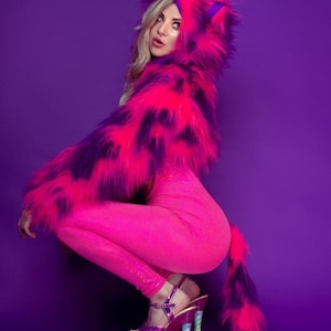 Può includere: Un crop top corto con cappuccio in pelliccia sintetica rosa e viola con una lunga coda. La modella indossa il top con leggings rosa e tacchi alti.