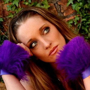 Peut inclure: Une femme aux longs cheveux bruns porte un pull en fourrure violet avec un ruban bleu. Elle regarde au loin avec une expression pensive.