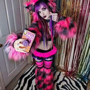 Puede incluir: Una mujer con un disfraz de gato de piel rosa y negra con polainas de piel rosa y negra. Est&aacute; sosteniendo un tel&eacute;fono con una funda de leopardo y un pop socket. El fondo es una cortina iridiscente arco&iacute;ris.