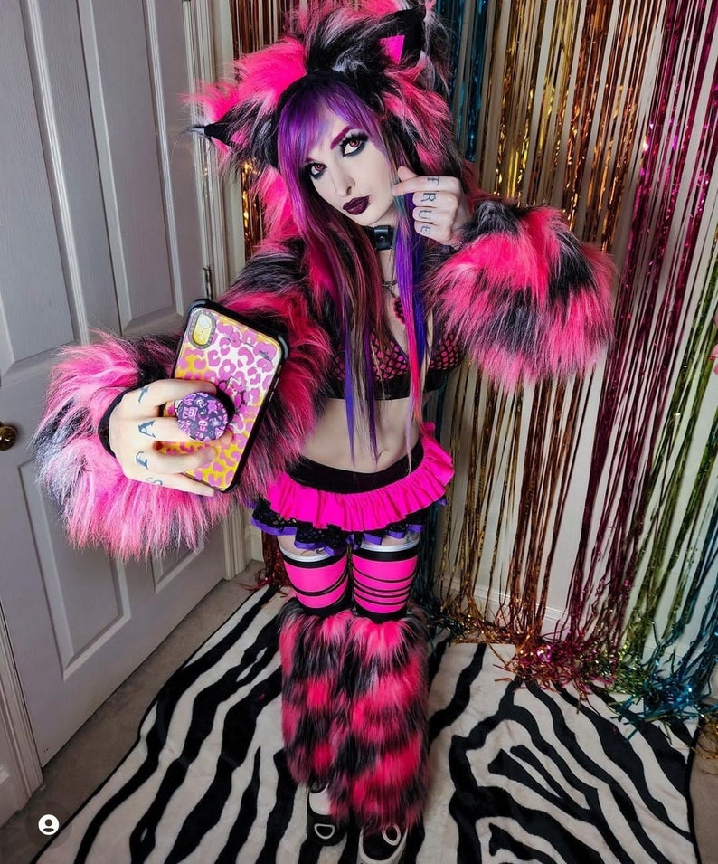 Puede incluir: Una mujer con un disfraz de gato de piel rosa y negra con polainas de piel rosa y negra. Est&aacute; sosteniendo un tel&eacute;fono con una funda de leopardo y un pop socket. El fondo es una cortina iridiscente arco&iacute;ris.