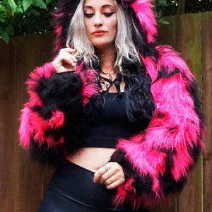Puede incluir: Una chaqueta de piel sintética rosa y negra con capucha de orejas de gato. La chaqueta es corta y tiene un corte holgado. La modelo lleva leggings negros y un top negro debajo de la chaqueta.